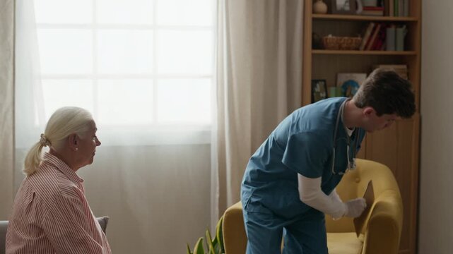 Young Caucasian male nurse examining senior Caucasian woman during home medical checkup, then sitting down and writing on clipboard. Ideal for healthcare services and geriatric care commercials