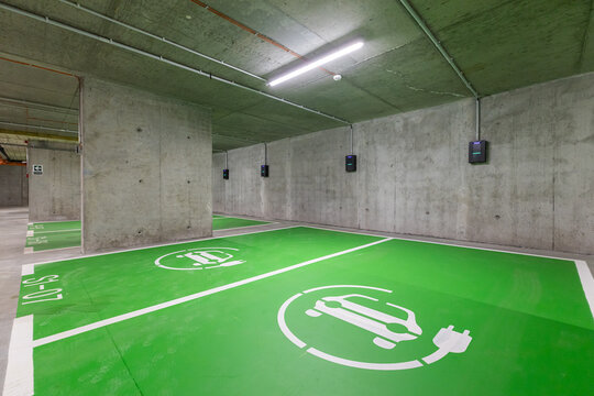 Electric cars parking lot. Modern underground concrete parking garage featuring bright green spaces marked with white electric vehicle charging symbols and wall-mounted EV chargers.