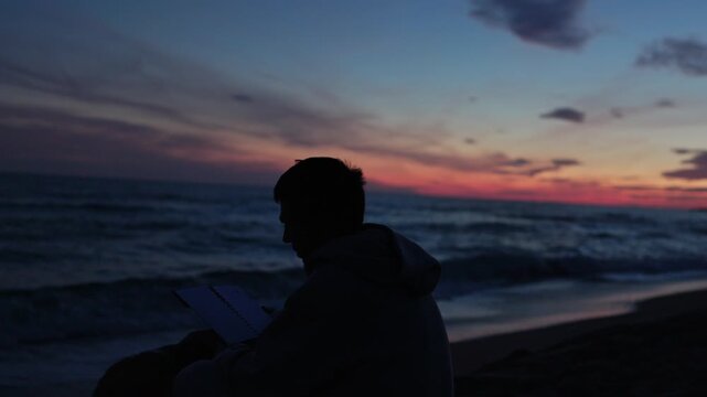 Silhouette of an inspired man sitting on a sandy beach at twilight, writing and drawing in his journal while watching the colorful sunset over the beautiful and calm ocean waves