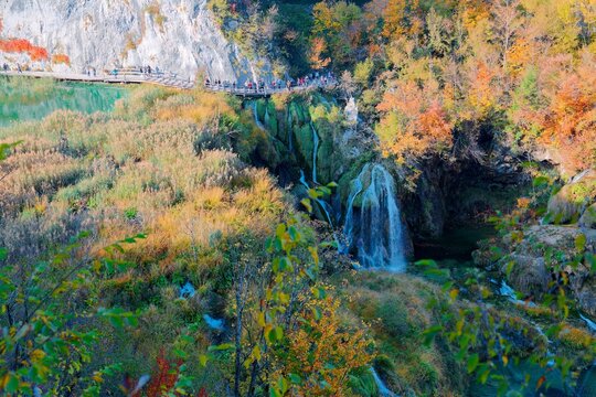 A high-angle aerial view captures the elegant Milanovac Waterfall cascading into Plitvice Lakes, flanked by golden autumn forests and a boardwalk bustling with tourists