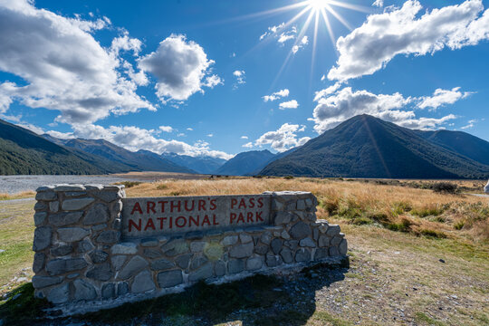 Steinernes Eingangsschild mit der Aufschrift ARTHURS Steinschild Arthurs Pass National Park vor Bergpanorama mit Sonnenstern in Neuseeland