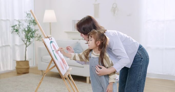 Adult asian single mother bends beside preschool daughter, guiding brush strokes on easel canvas in home art lesson, weekend bonding time, early childhood development, cozy room, gentle parenting.