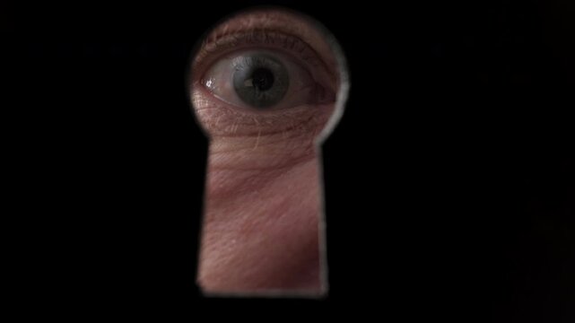 Person's eye looking through keyhole on black door close-up