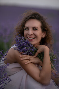 Joyful young woman holding fragrant Lavandula bouquet while sitting in a purple field during a scenic summer sunset