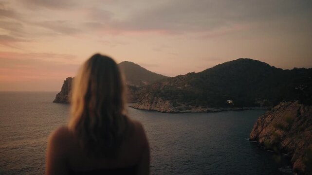 A young woman meditates quietly on a rocky Ibiza coastline, gazing at the glowing sunset over tranquil sea and dramatic hills. Warm golden hour lighting sets a peaceful, contemplative mood.