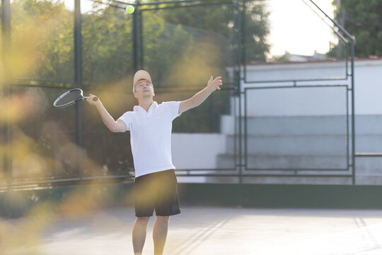 Man serves a tennis ball on a court during late afternoon practice session in a park