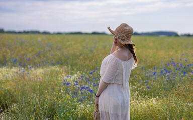 Woman Looking into the Distance in a White Dress and Straw Hat. A woman in a flowing white dress and straw hat stands with her back partially turned toward the camera, gazing across a vast summer mead © Nadiia