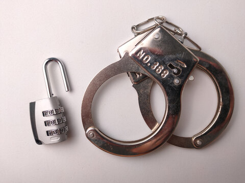 An open silver combination padlock placed next to shiny metal handcuffs on a plain white surface