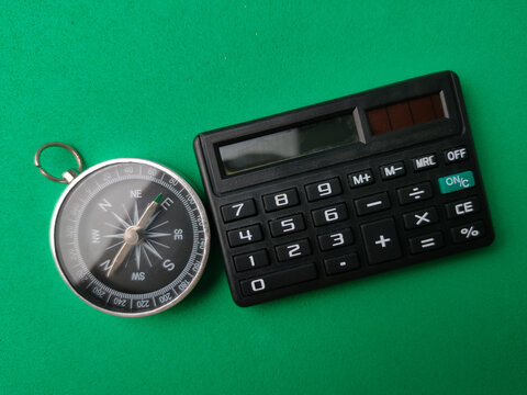 A metallic compass placed alongside a compact black pocket calculator on a smooth green background