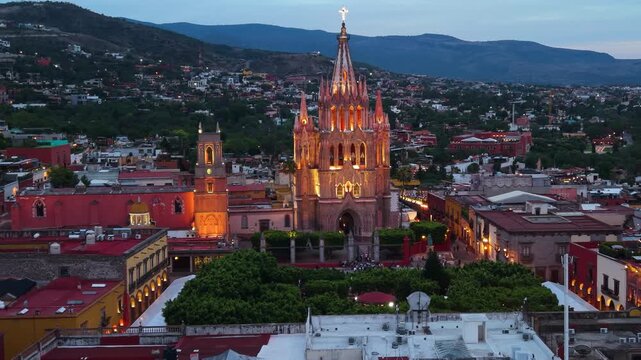 A cinematic twilight aerial view capturing the illuminated neo-Gothic facade and spires of the iconic Parroquia de San Miguel Arc&aacute;ngel.
