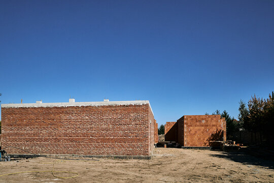 Wide horizontal view of a residential building construction site featuring red brick and hollow ceramic block walls with a reinforced concrete ring beam under a clear blue sky.