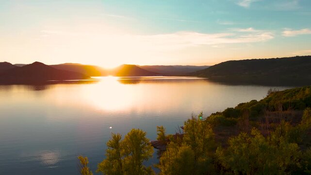 A drone shot of a magnificent sunset over a lake behind some hills, with the reflection on the water, vegetation in the foreground, and a clear blue sky