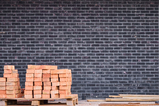 Stacked red clay bricks on a wooden pallet in front of a dark clinker brick wall with copy space at a construction site.