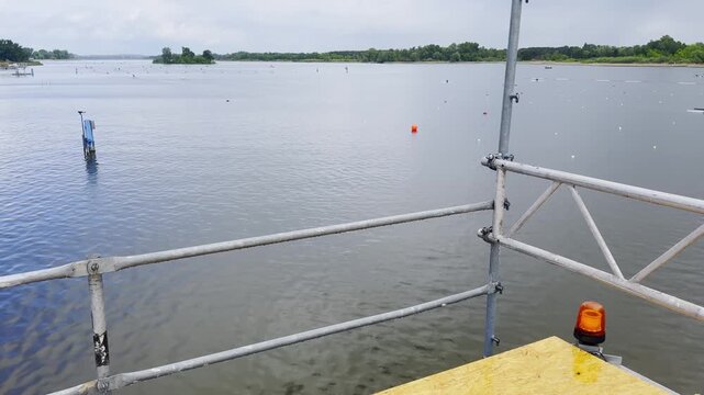 Floating event platform on a lake with metal scaffolding railings, plywood deck, orange warning beacon, cables and calm water in the background.