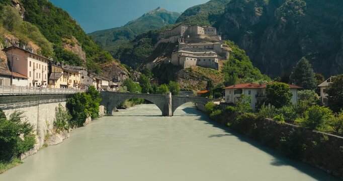 Cinematic drone flight above a glacial river toward an arched bridge, revealing historic Fort Bard, colorful village houses and towering Alpine peaks in Italy's Aosta Valley.