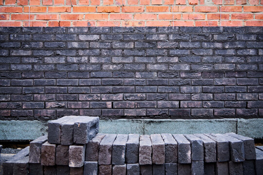 Front view of a dark clinker brick wall under construction with a stack of loose facing bricks in the foreground and a red brick backing wall.