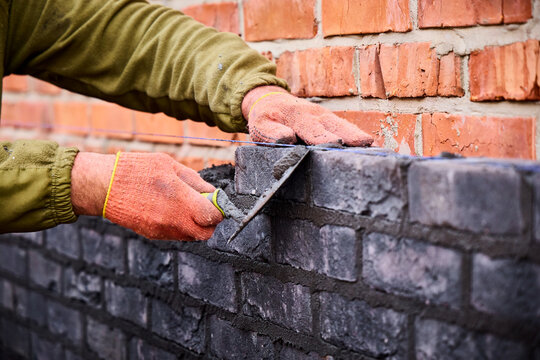 Close-up of a bricklayer's hands in orange protective gloves using a trowel to align a dark clinker brick along a blue mason line during wall construction.