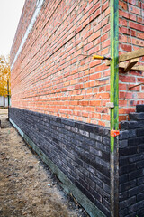 Low-angle perspective view of a long exterior wall under construction, featuring dark clinker brick facing along the base, red brick backing masonry with wall ties, and a green corner alignment profil © Oleg