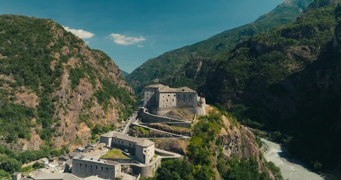 Drone rises above historic Fort Bard, revealing the winding river and deep Aosta Valley framed by lush green mountains beneath a clear blue summer sky in northern Italy.
