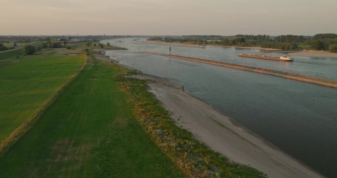 Drone approaches a cargo ship from the riverbank, revealing a winding river, protective dikes and the expansive Dutch polder landscape, showcasing inland shipping and water management.
