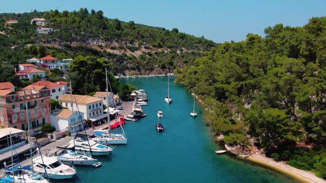 Aerial tracking shot flying down the narrow turquoise Ionian sea channel of Gaios port in Paxos, Greece, flanked by yachts and a green nature under bright summer sky
