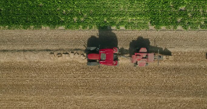 Slow-motion minimalistic shot of a red tractor baling straw from harvested wheat chaff, casting a striking shadow across the field under the high summer sun in a cinematic rural landscape.