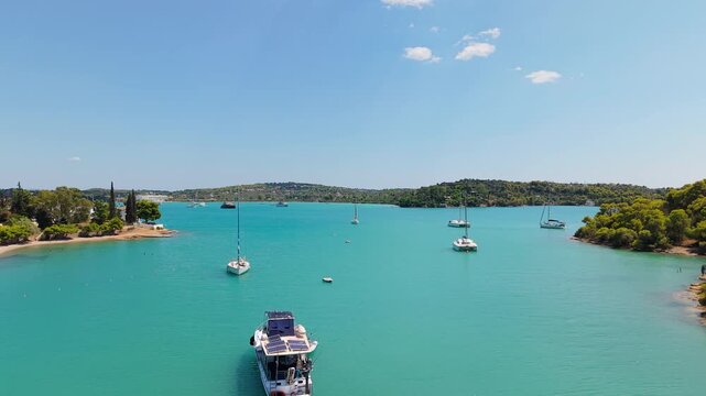 Drone flies forward over Porto Cheli's stunning turquoise bay in summer. Sailboats and yachts drift peacefully between lush green peninsulas under a bright midday sun.