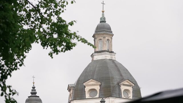 Wide angle footage shows the iconic dome of Pažaislis Monastery in Kaunas, Lithuania. Overcast skies and leafy branches frame baroque architecture in soft natural daylight. No people visible.