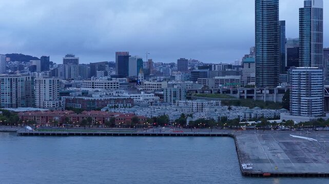 Drone view above San Francisco&rsquo;s Embarcadero waterfront, revealing sleek high-rises, city skyline, and pier in moody evening light with dramatic clouds overhead.