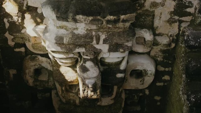 Detailed close-up shot of the weathered Chaac mask carved in stone at Tikal, Guatemala. Soft sunlight highlights textures of the famous Mayan ruins, revealing sacred artistry.