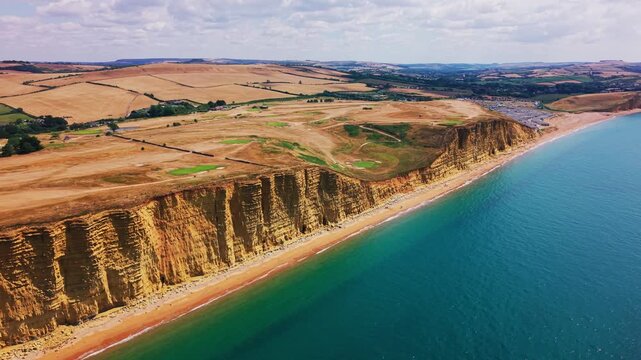 Sweeping aerial drone footage of the golden cliffs, turquoise sea and rolling countryside at West Bay on Dorset&rsquo;s Jurassic Coast, England. The wide coastal view reveals layered rock formations