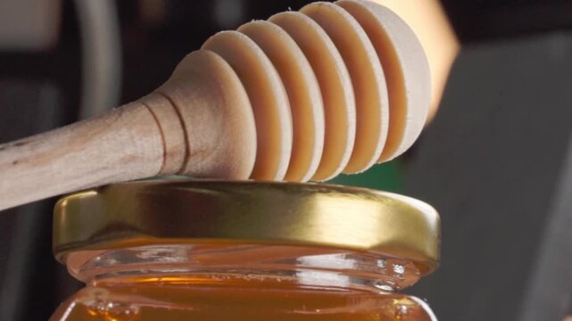Close up panning shot of a wooden honey dipper resting on a glass jar filled with golden honey, highlighting natural sweetness and beekeeping products
