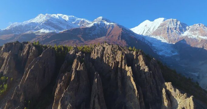Drone gliding shot over a pine tree-covered eroded cliff ridge on Annapurna Circuit, Manang Nepal, with snow-capped Annapurna mountains in the background