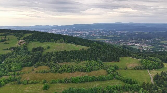 Expansive rural landscape with emerald hills, patterned fields, wooded slopes, winding roads, and faraway settlement beneath soft overcast light, creating calm natural scenery with broad depth and