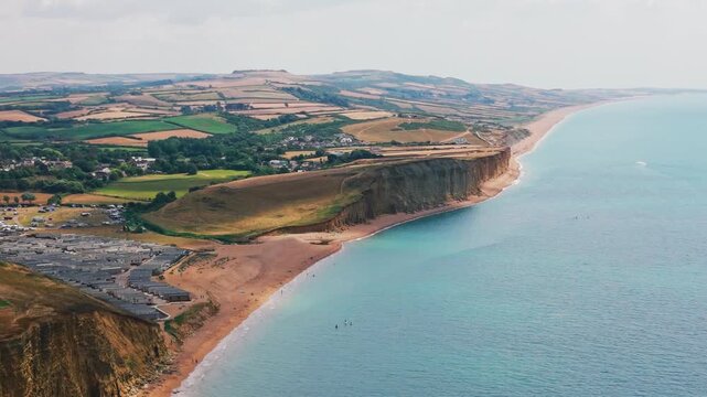 Sweeping aerial drone footage looking east from West Bay across dramatic coastal cliffs, beaches and patchwork farmland toward Burton Bradstock in Dorset, England. Wide summer landscape featuring calm