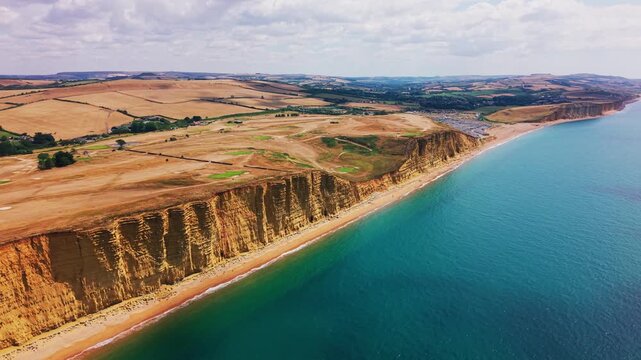 Wide drone footage looking along the dramatic golden cliffs, turquoise sea and rolling farmland near West Bay on Dorset&rsquo;s Jurassic Coast, England. Expansive coastal landscape showing a narrow beach
