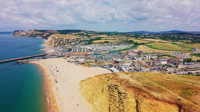 Sweeping aerial drone footage overlooking West Bay&rsquo;s shingle beach, harbour, coastal town and golden East Cliff on Dorset&rsquo;s Jurassic Coast, England. Wide summer establishing shot featuring blue sea