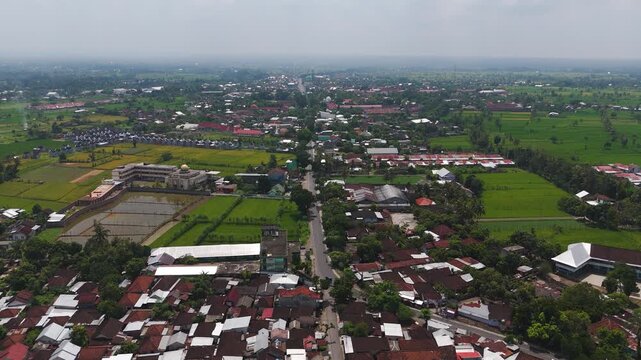 Straight paved road runs through dense red tile rooftops in Pelowok Selatan, Kediri, West Lombok Regency, gold domed mosque and green rice fields spread into distance. Aerial drone view.