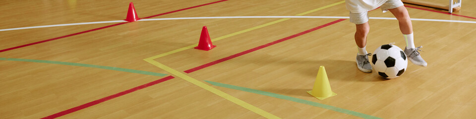 Fototapeta premium Header cropped shot of child practicing soccer dribbling around cones on indoor gym floor. Image suitable for youth sports programs, physical education, and kids fitness marketing