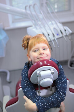 A happy red-haired girl hugs the dentist's chair, smiling in the doctor's office. Positive pediatric dentistry. A successful dentist visit without fear.