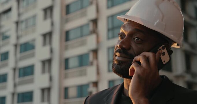 African American construction engineer in a white hardhat talks on a smartphone outside a building, coordinating contractors, partners and project decisions.