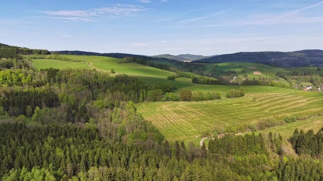 High viewpoint reveals rolling green hills and patchwork meadows bordered by dense forests, with scattered village homes across wide rural countryside beneath clear blue sky in bright daylight
