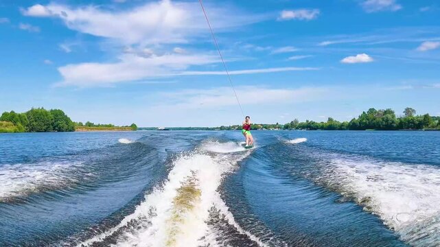 woman wakeboarding behind boat at high speed holding tow rope on wide river. athletic female glides fast on water surface creating wake. wakeboarding extreme water sport and active summer adventure