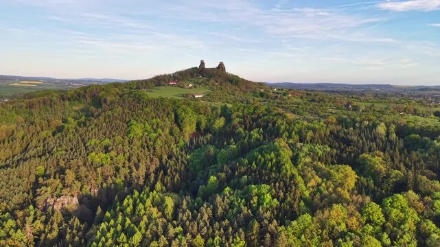 Ancient stone castle ruins stand on grassy hill above dense woodland, scattered cottages, and patchwork farmland, with clear sky, warm daylight, and distant valley creating calm rural scenery