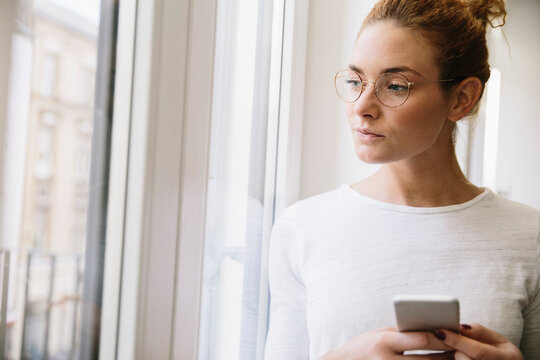 Young woman looking up from her mobile phone, out of window