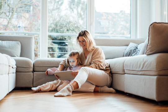 Mother sharing tablet PC with daughter sitting by sofa in living room