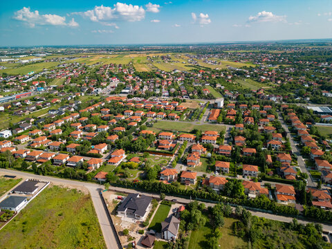 High angle drone shot showing vibrant residential neighborhood with suburban houses, red roofs, green trees, and blue sunny sky in Domnesti, Ilfov, Romania.