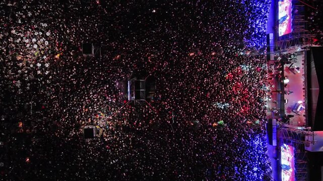 Aerial top view of crowd at night music concert. People gathered at festival outdoor stage with lights and screens, 4k