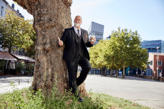 Elegant businessman leaning on tree in the city, drinking coffee