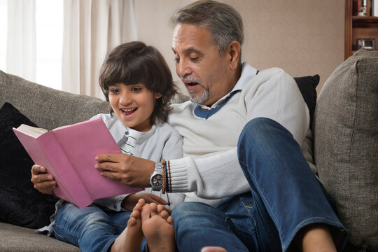 Grandfather and grandson reading book sitting together on sofa
